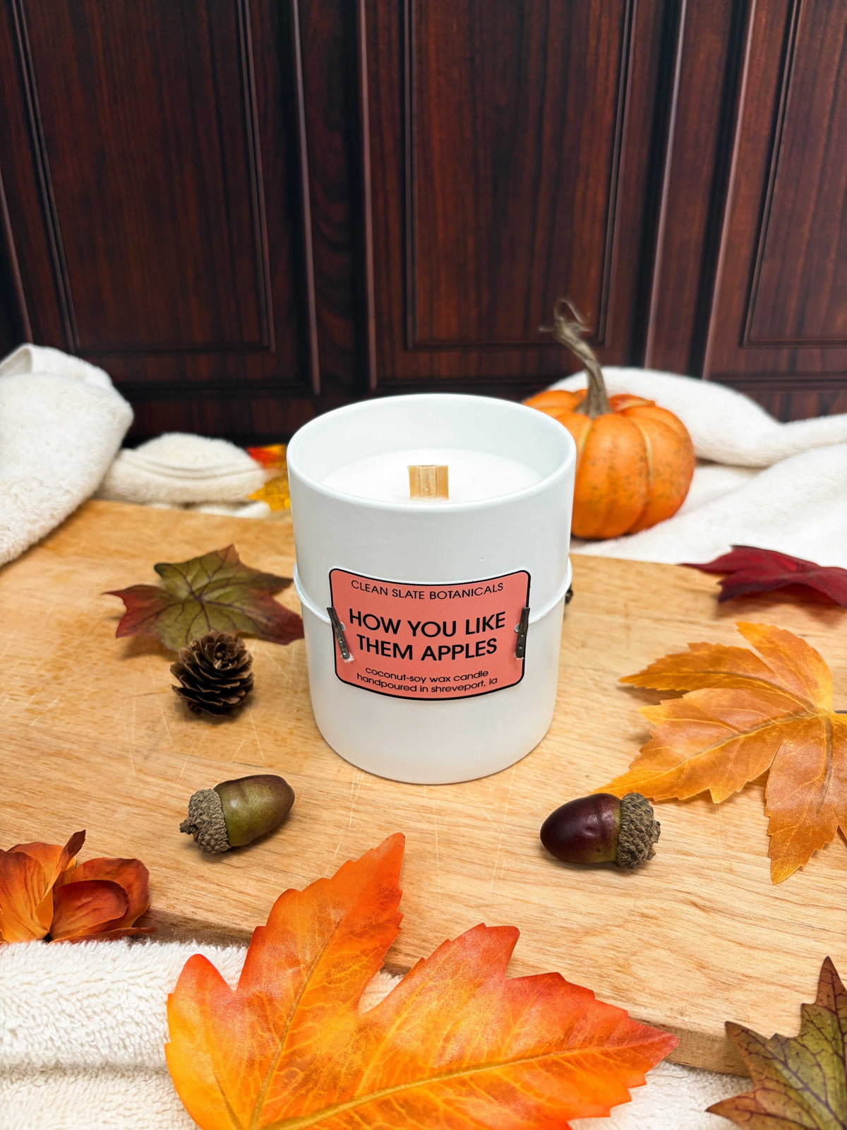 How You Like Them Apples candle in a white ceramic jar with a coral label and wooden wick, surrounded by autumn leaves