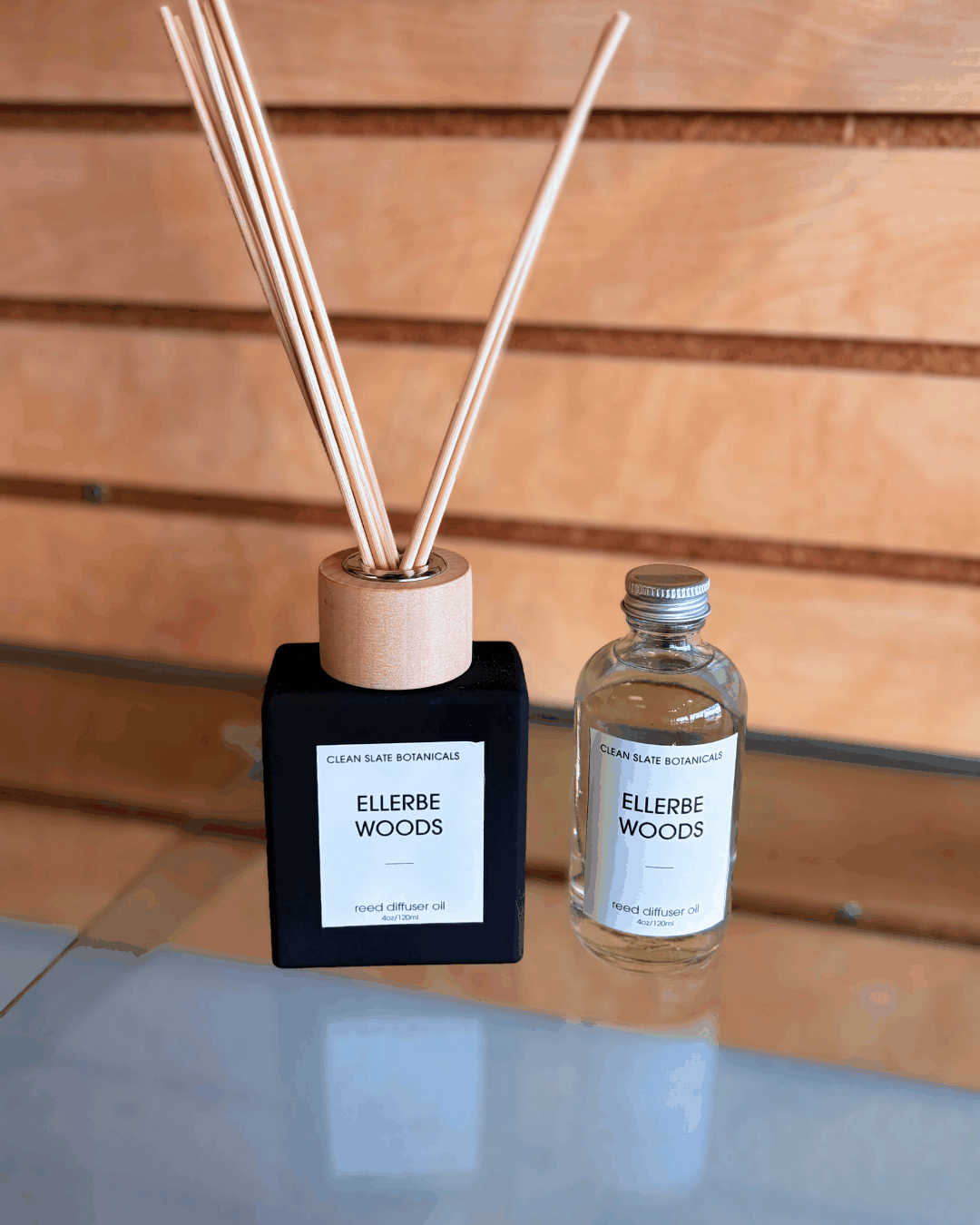 Ellerbe Woods Reed Diffuser in a black square glass bottle with pale reeds, beside a clear refill bottle