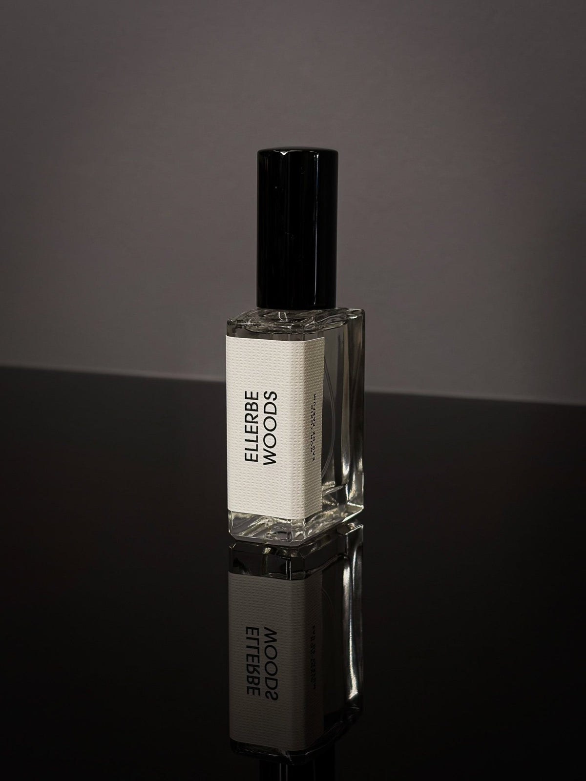 Ellerbe Woods Wearable Fragrance clear rectangular glass bottle with black cap and white label on a reflective black surface