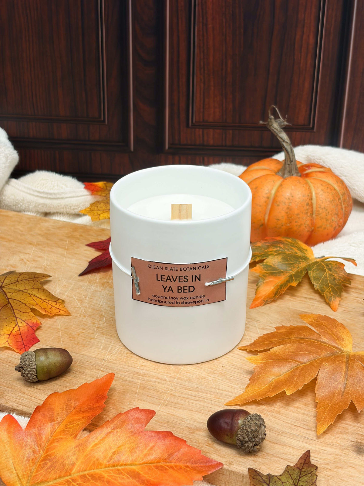 Leaves In Ya Bed Candle in a white glass jar with a wooden wick and brown label, surrounded by autumn leaves