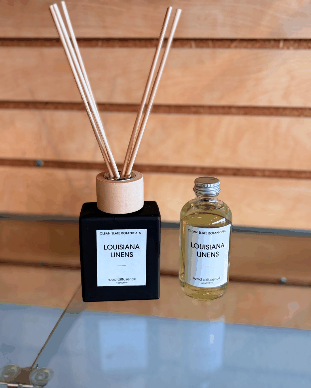 Louisiana Linens Reed Diffuser with black square base, natural wooden reeds and a clear refill oil bottle on glass surface
