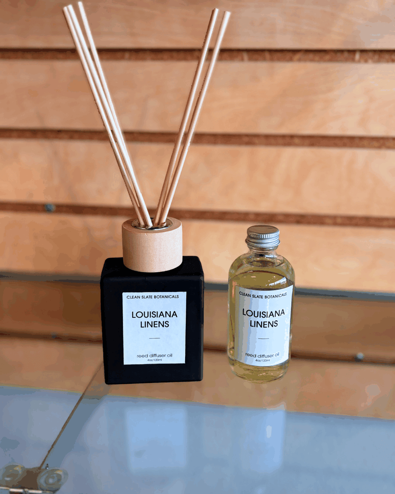 Louisiana Linens Reed Diffuser with black square base, natural wooden reeds and a clear refill oil bottle on glass surface