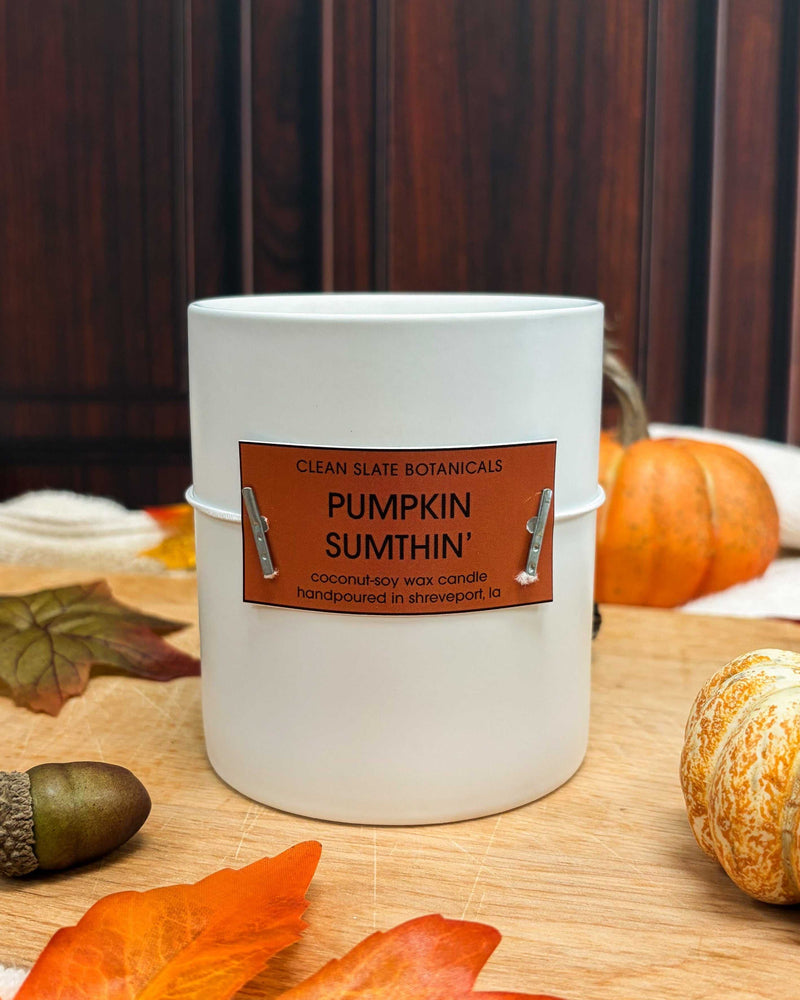 Pumpkin' Sumthin' Candle in a white vessel with an orange label, beside miniature pumpkins and autumn leaves