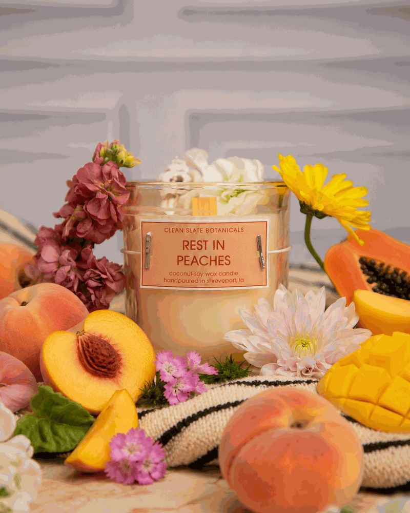 Rest In Peaches candle in a clear glass jar with a peach label, surrounded by fresh peaches, mango, and colorful flowers