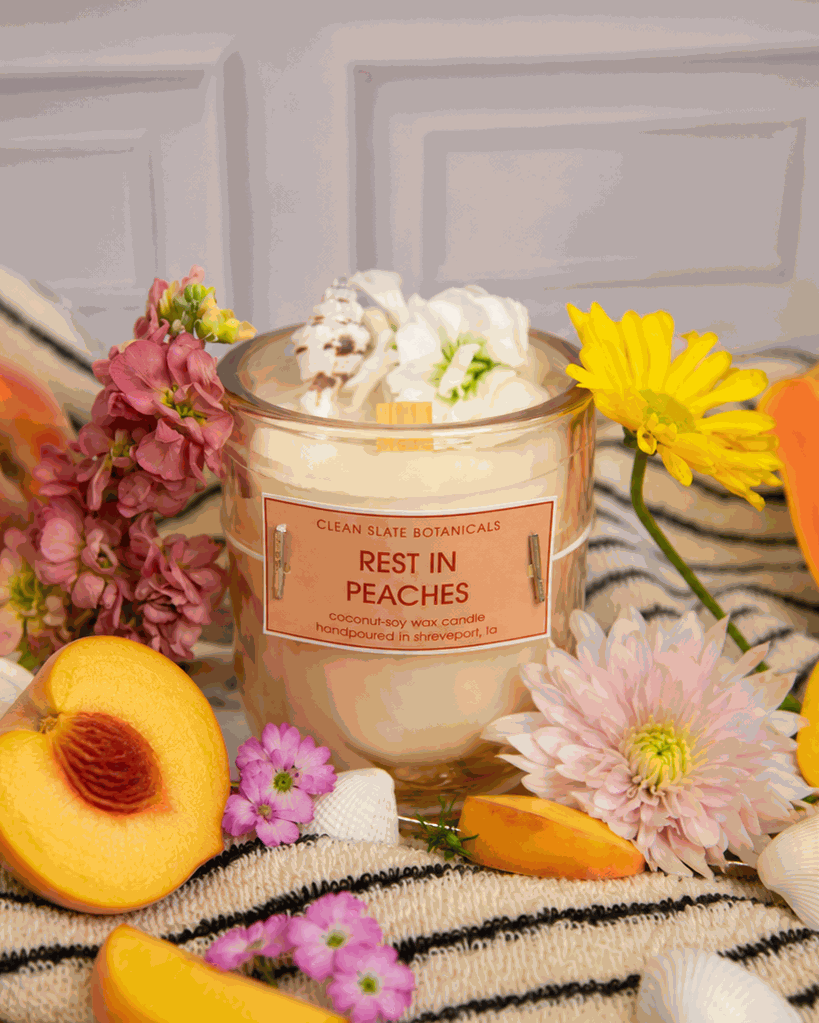 Rest In Peaches candle in a clear glass jar with cream wax and a peach label, surrounded by flowers and peach slices