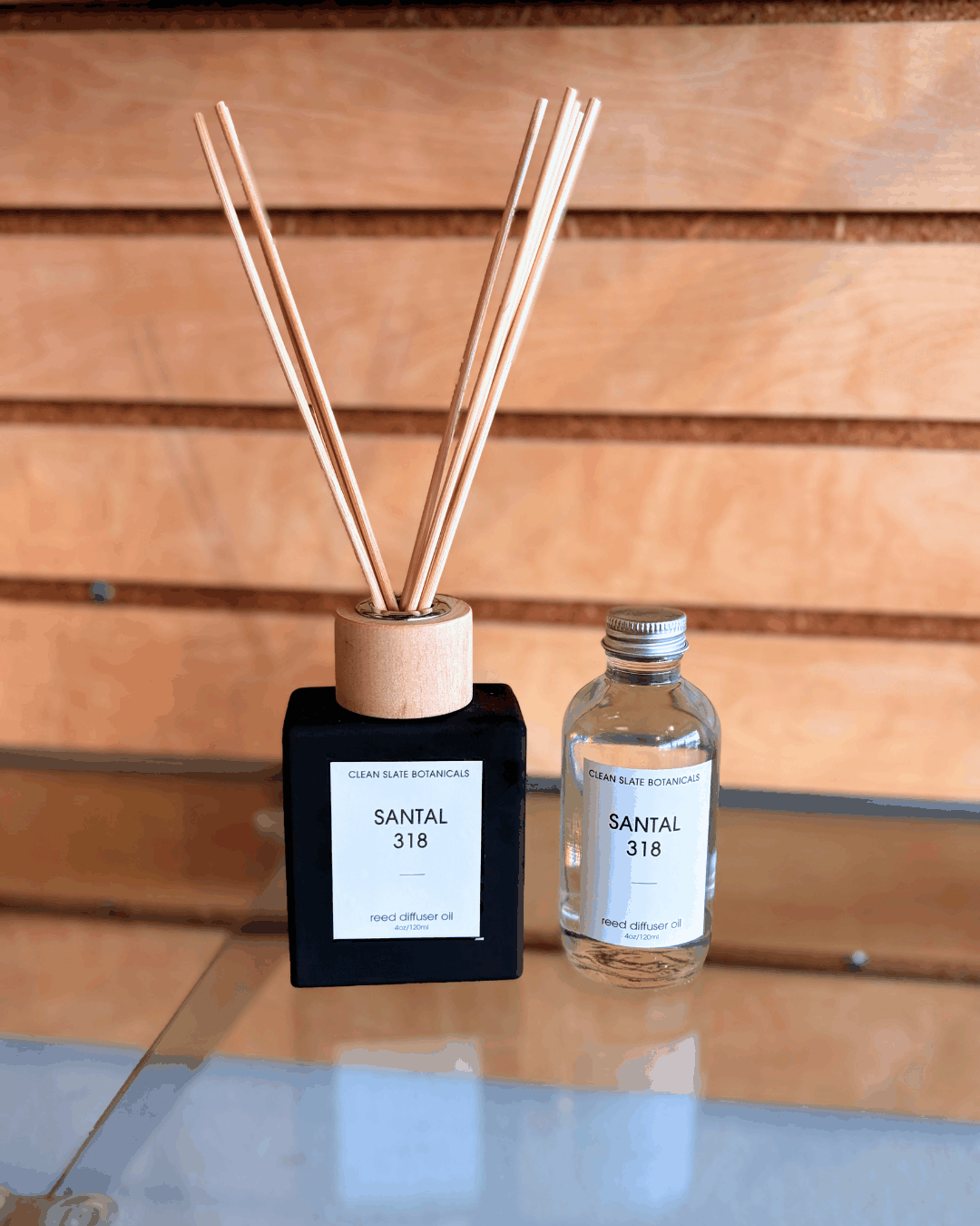 Santal 318 Reed Diffuser in a black square bottle with a pale wood collar and reeds, beside a clear glass refill bottle