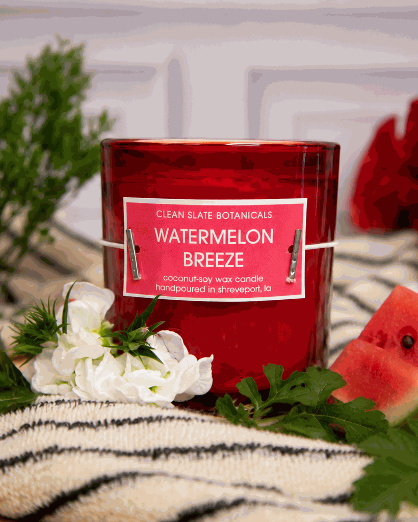 Watermelon Breeze Candle in a translucent red glass jar with a red label and white lettering, beside watermelon